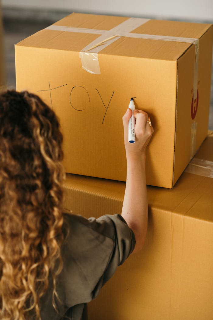 Back view of crop faceless female in casual wear labeling packed cardboard box with marker while moving house