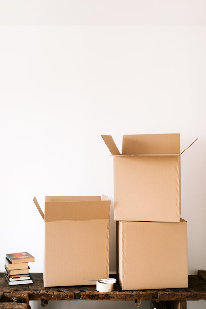 Unsealed carton boxes placed on shabby wooden table near stack o paper books and tape against white wall in daylight